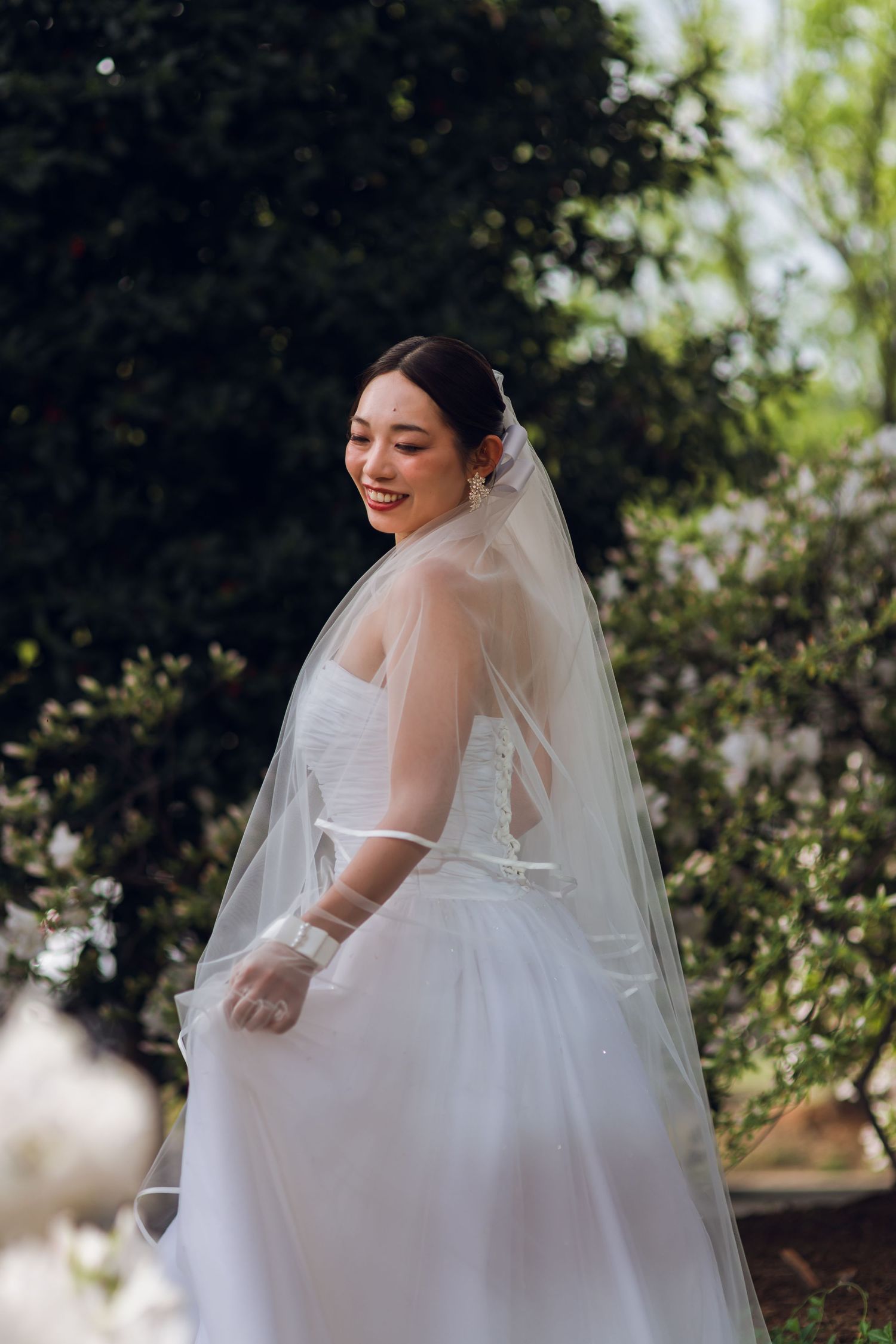 Smiling Asian bride in white wedding gown and veil poses outdoors in a garden setting.