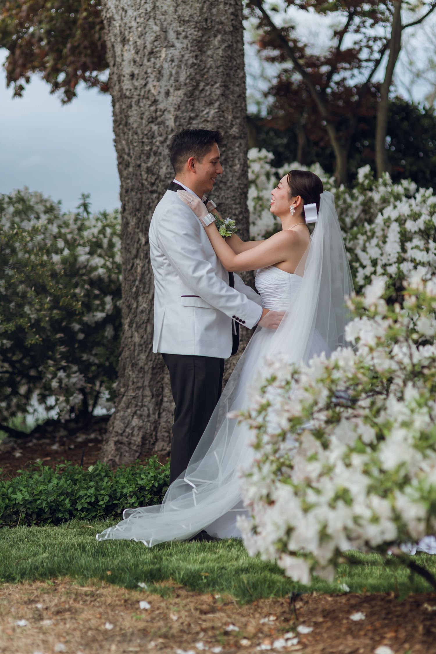 A bride and groom share a romantic embrace near a tree surrounded by white blooming flowers outdoors.