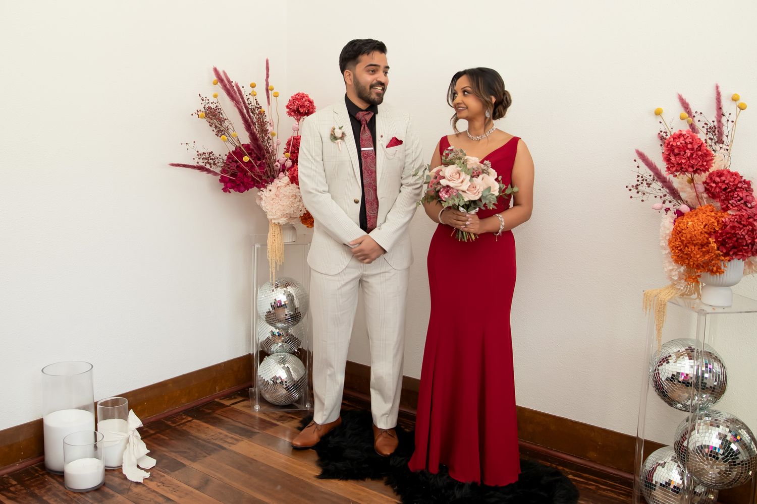 Couple in elegant wedding attire, man in white suit and woman in red gown, standing before floral arrangements.