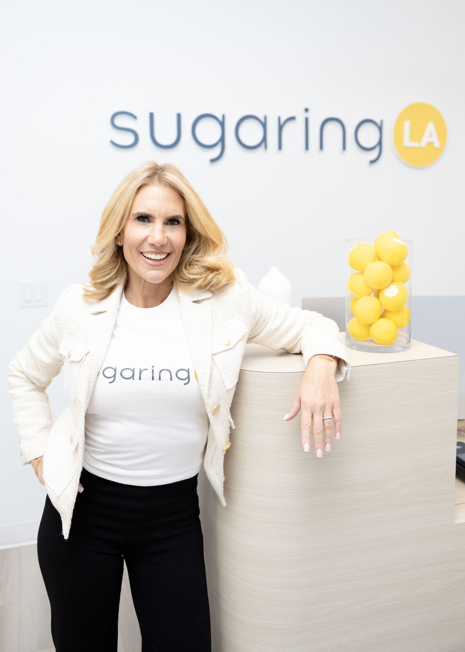 Woman in white Sugaring LA shirt smiles at reception desk with lemon decor and Sugaring LA logo on wall.