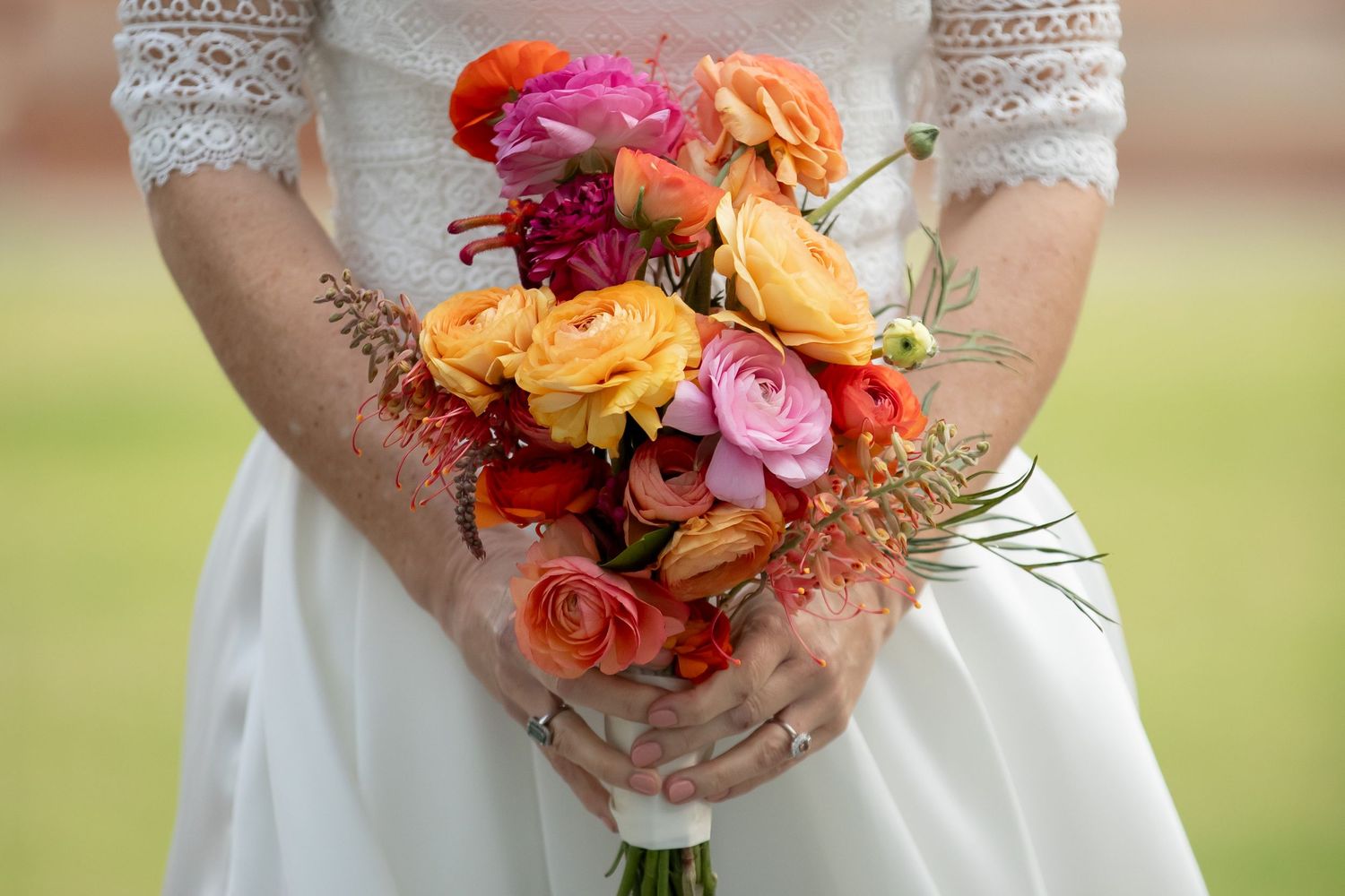 Bride holding a vibrant bridal bouquet of orange, pink, and red roses and ranunculus in a white lace wedding dress.