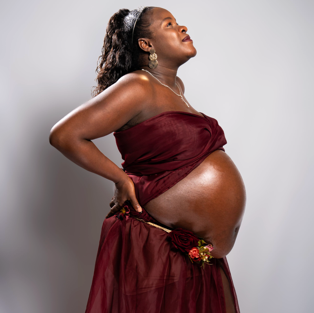 Pregnant woman in elegant burgundy draped gown posing gracefully with floral accent, maternity photography studio portrait.