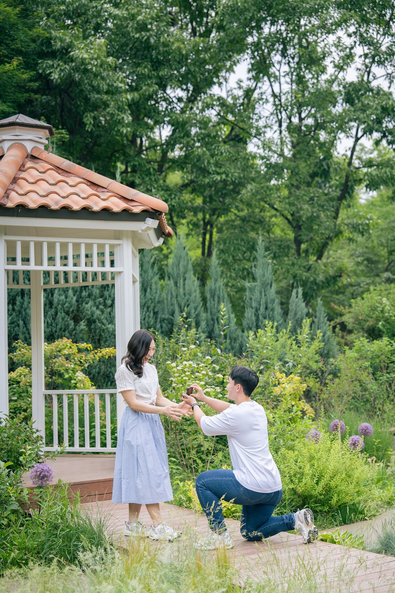 Man proposes to woman on one knee in a lush garden near a white gazebo with terracotta roof.