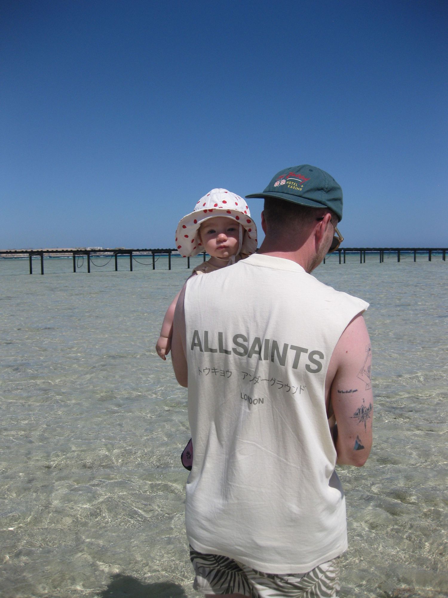 Father holding baby on his hip while standing in clear shallow ocean water at a sunny beach.