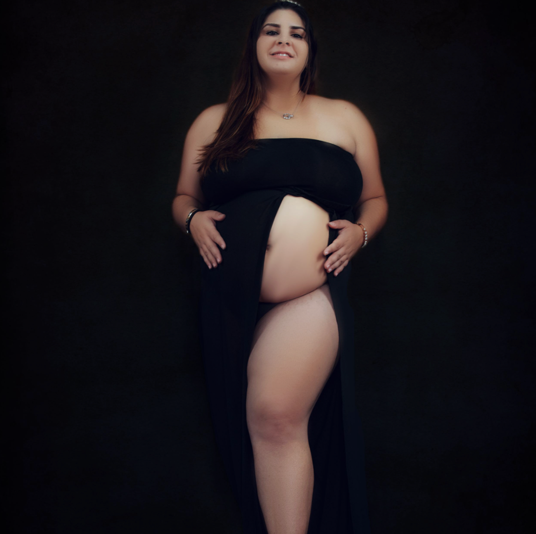 Pregnant woman in black cutout maxi dress poses against white backdrop in elegant maternity photo shoot.