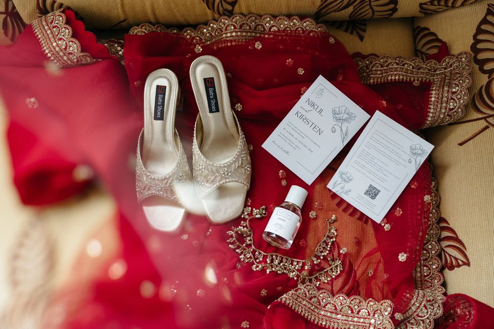 Wedding flat lay with embellished heels, red bridal lehenga, jewelry, and invitation cards.