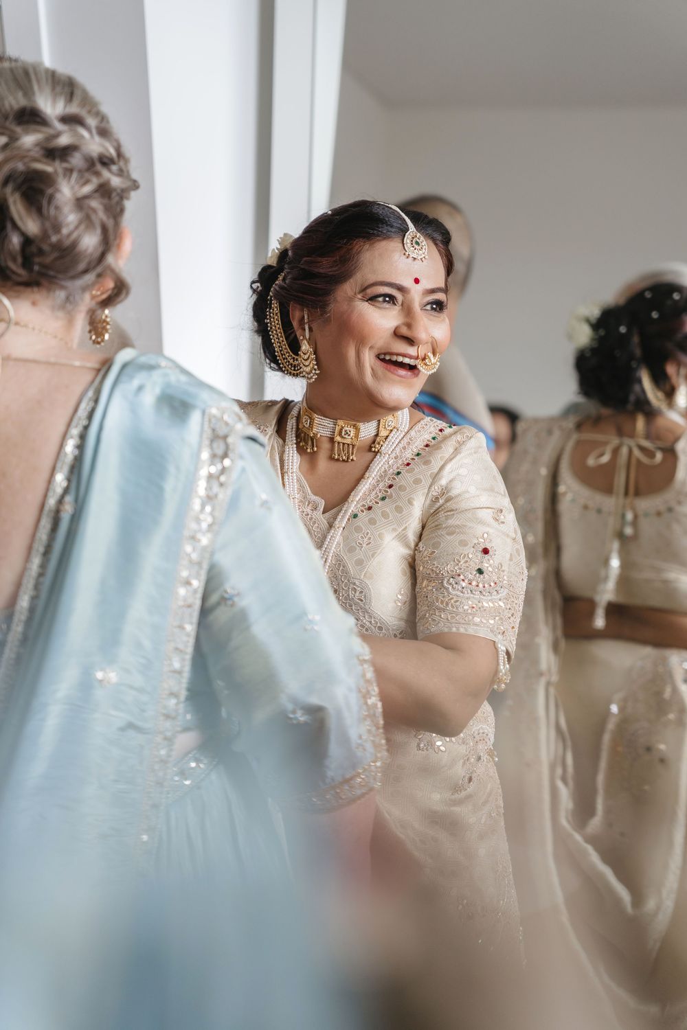 Smiling Indian bride in white saree with gold jewelry and maang tikka surrounded by guests at wedding celebration.