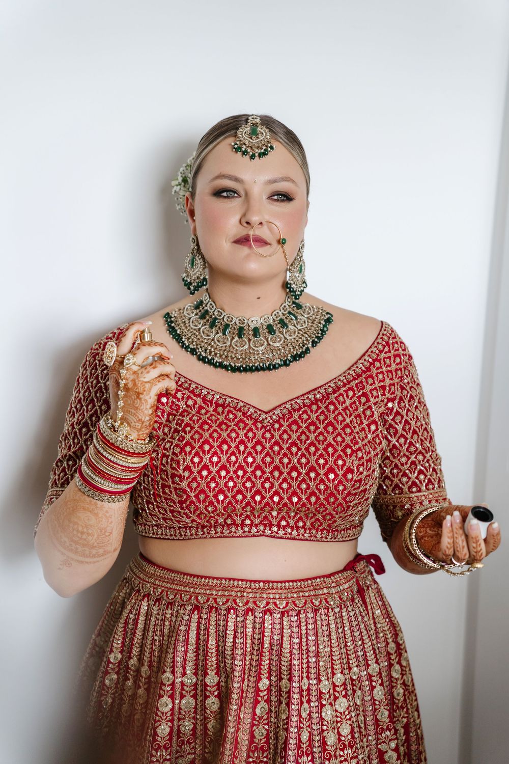 Bride in traditional red Indian lehenga with gold jewelry and mehndi posing indoors.