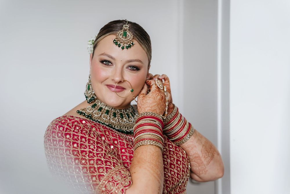 Bride in red embroidered lehenga with green gold jewelry and mehndi adjusting earring on wedding day.
