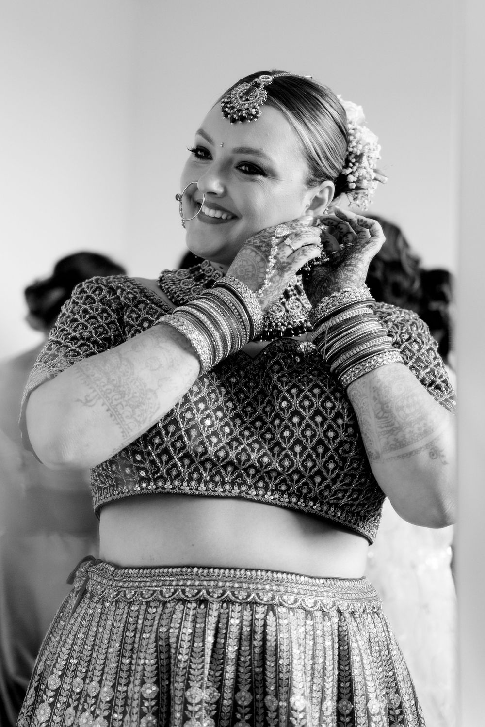 Black and white photo of a smiling bride in Indian lehenga, maang tikka, bangles, and mehndi adjusting her earrings.