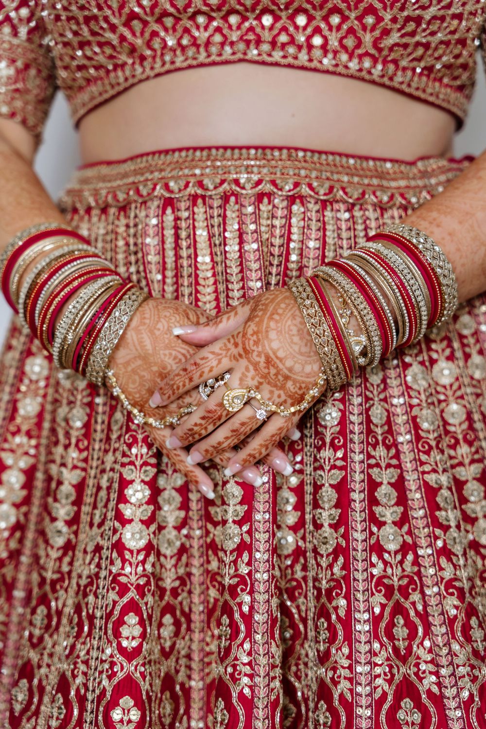 Indian bride's hands adorned with intricate mehndi henna and gold bangles against a red and gold embroidered lehenga.