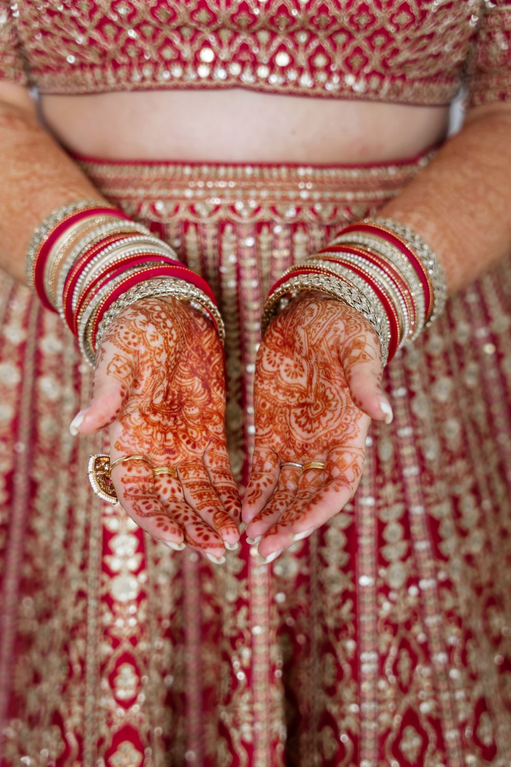 Close-up of Indian bride's palms showing detailed mehndi henna art, wearing red and gold bangles with embroidered lehenga.