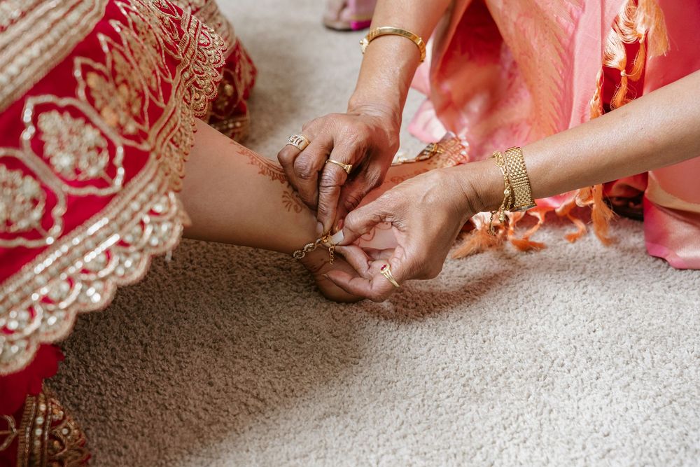 Close-up of hands adorned with gold bangles and henna adjusting bridal ankle jewelry on a red embroidered sari.