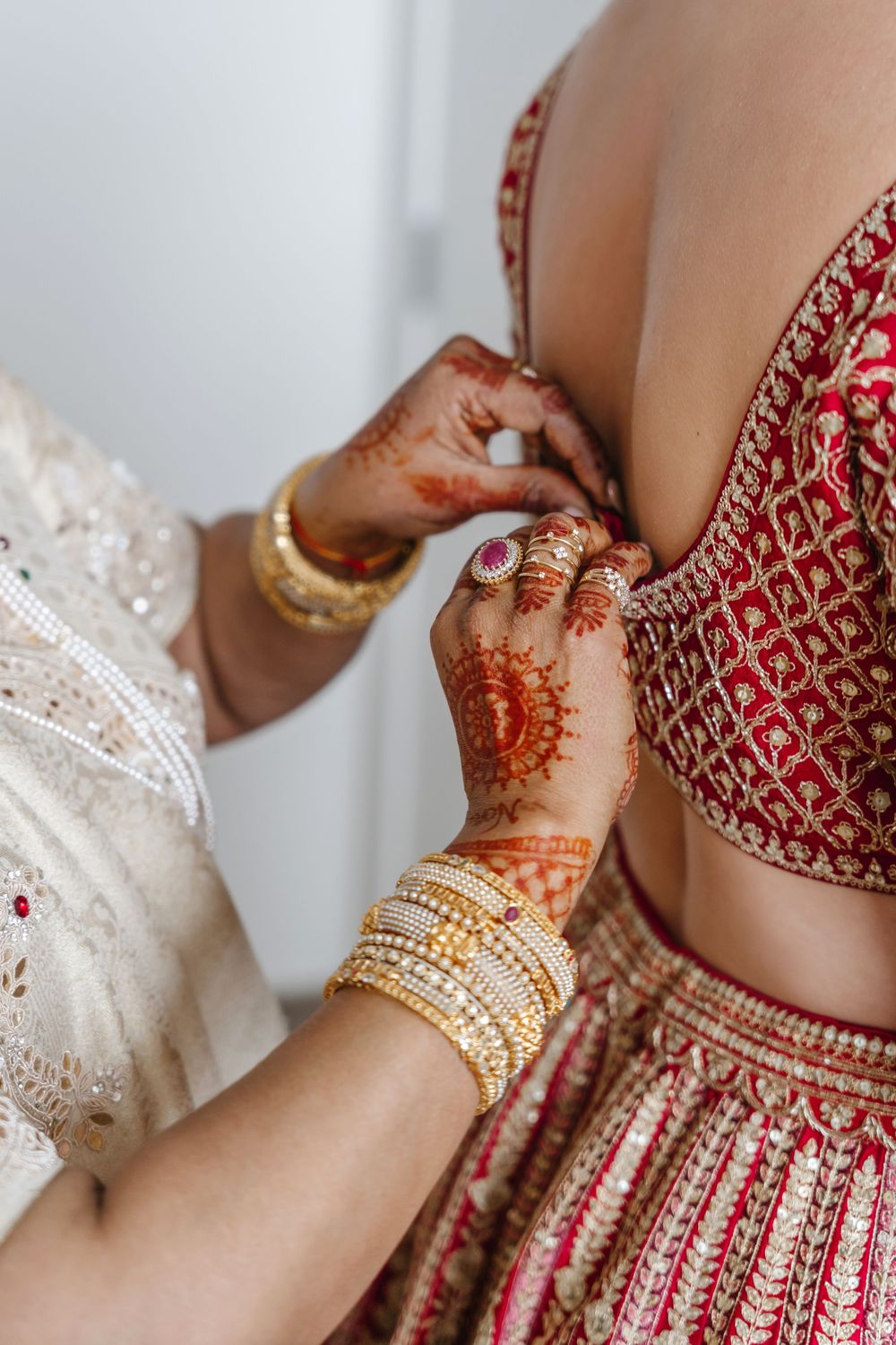 Hands adorned with henna and gold bangles fastening a red and gold bridal lehenga blouse.