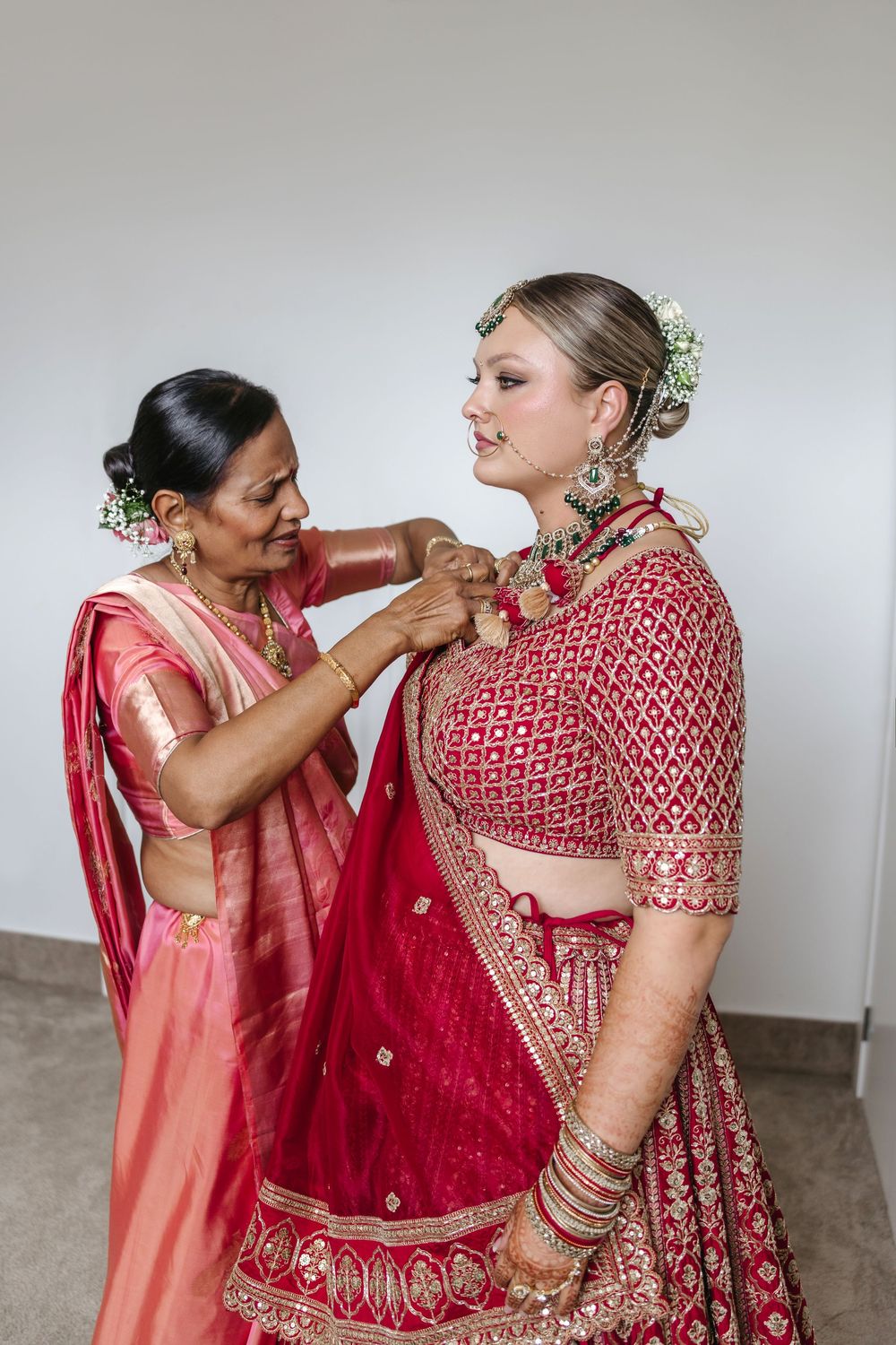 Two women in red Indian bridal attire, one adjusting the other's dupatta, adorned with traditional jewelry.