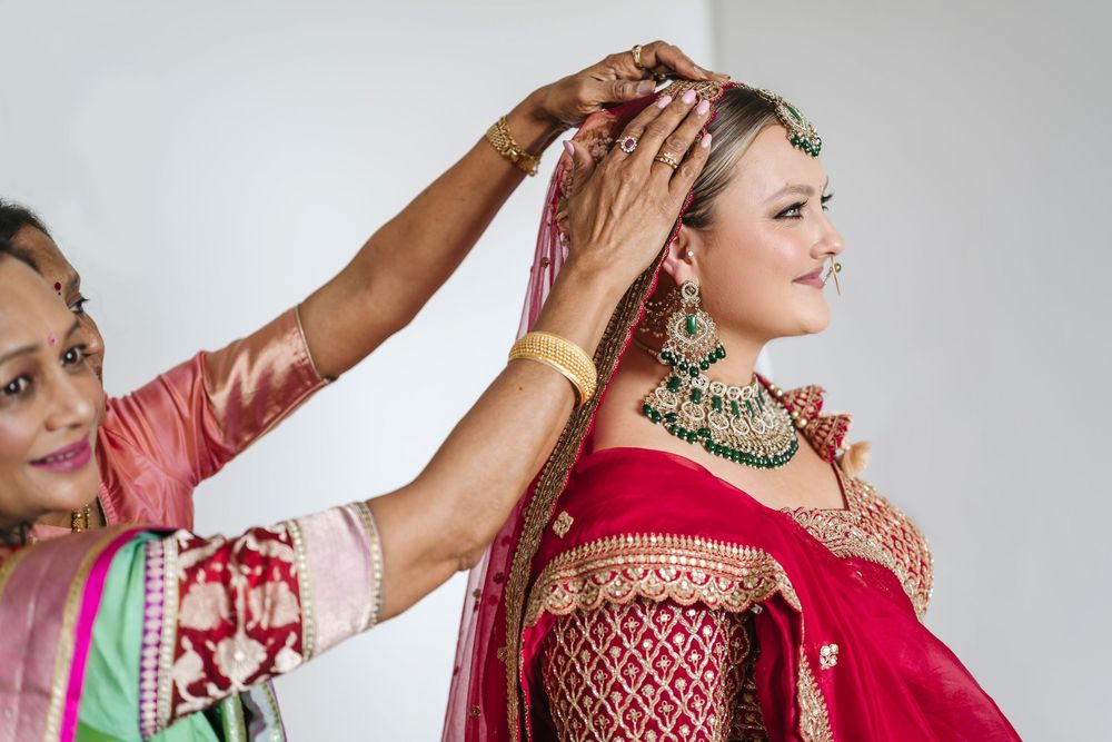 A woman in red bridal lehenga gets dupatta arranged by helper during Indian wedding preparation.