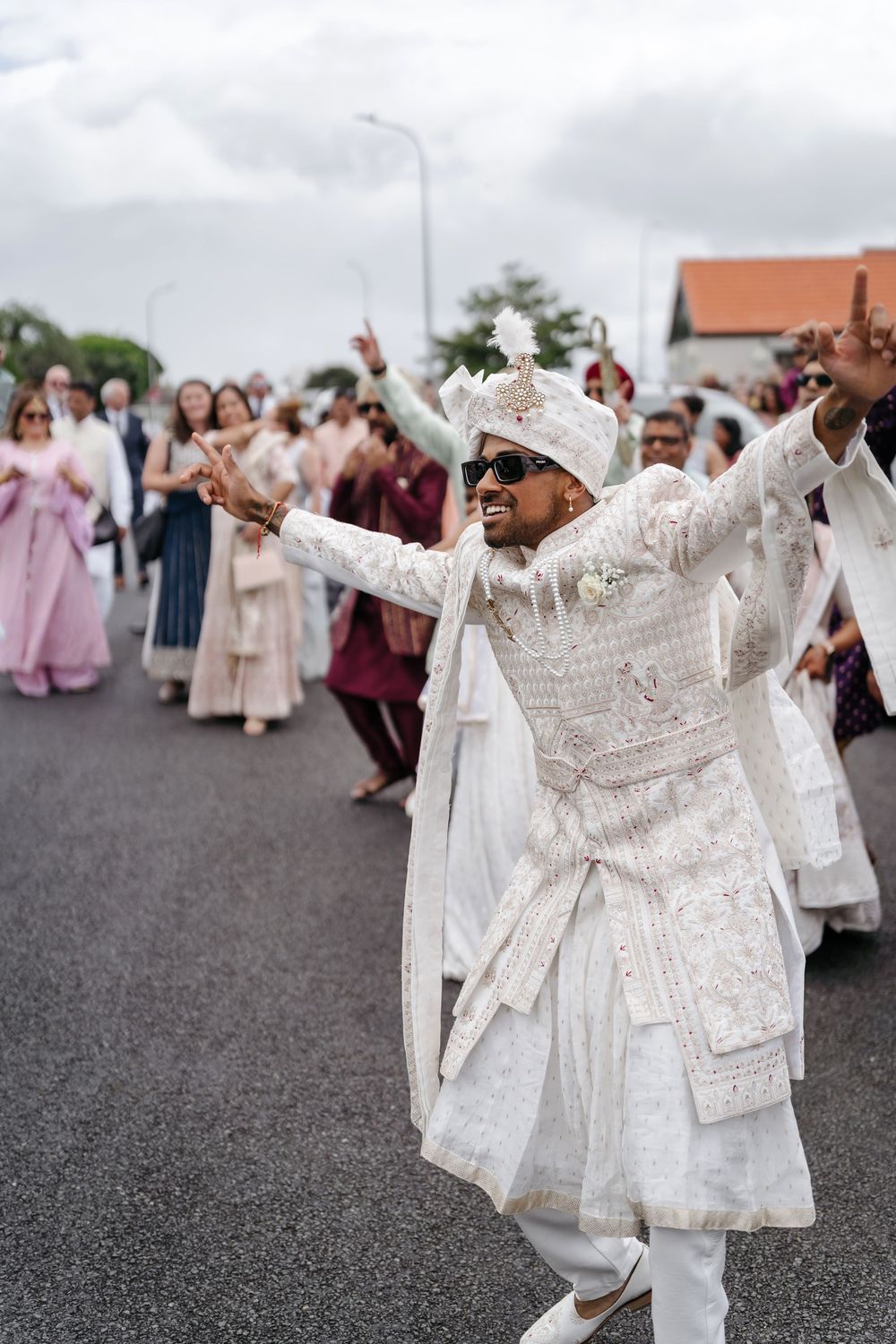 Indian groom in white sherwani and turban dancing joyfully during a traditional Baraat wedding procession outdoors.