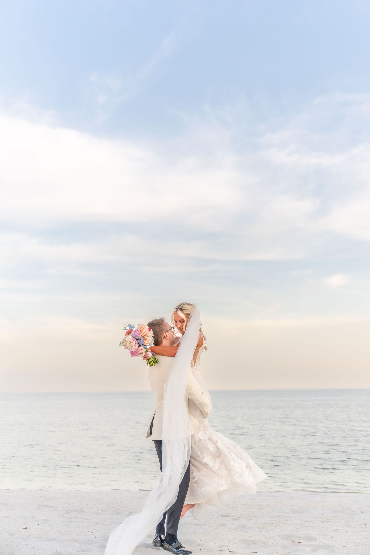 Groom lifting bride in wedding dress on beach with bouquet, ocean backdrop at dusk.