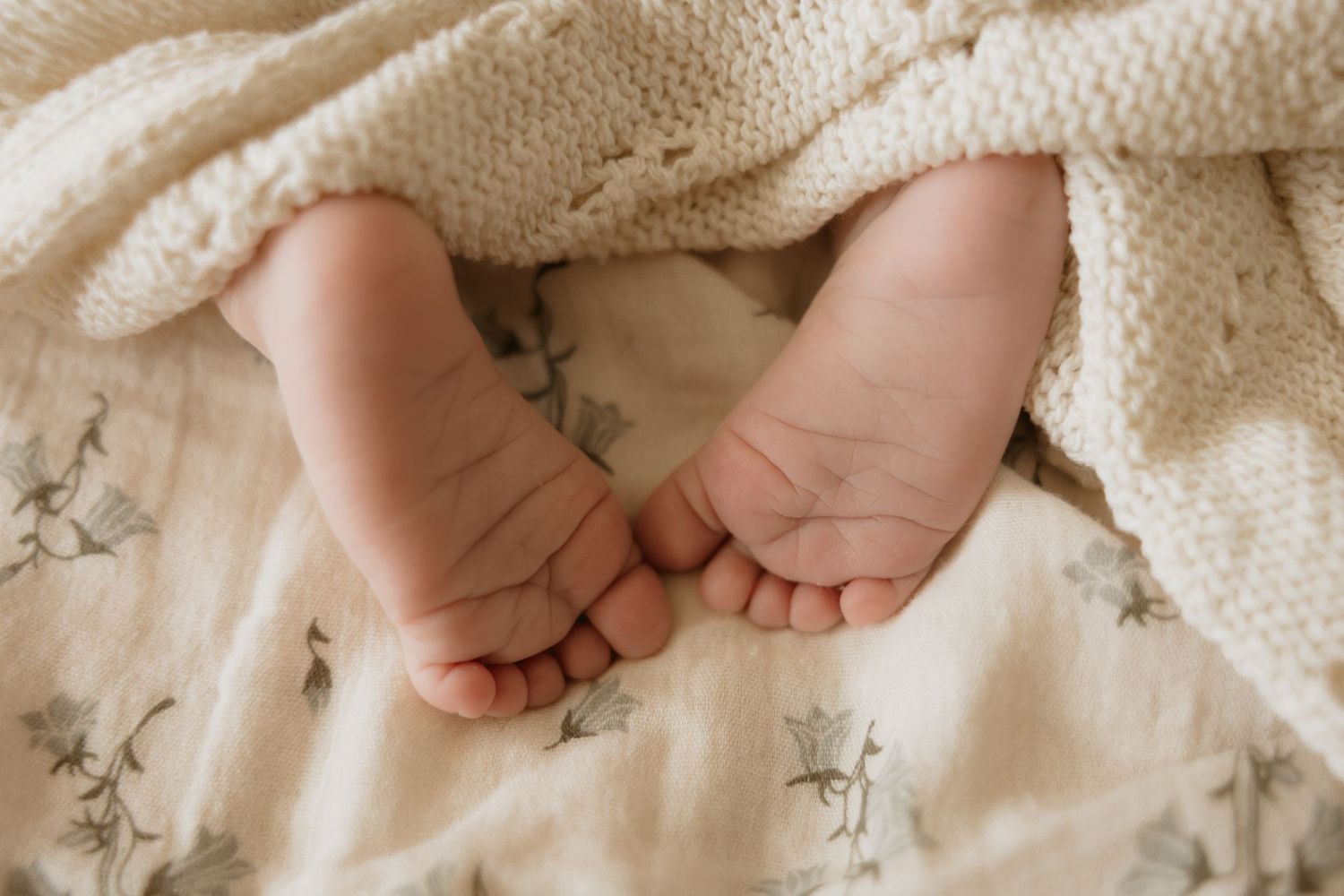 Close-up of tiny newborn baby feet peeking out from a soft cream knit blanket on a floral muslin surface.