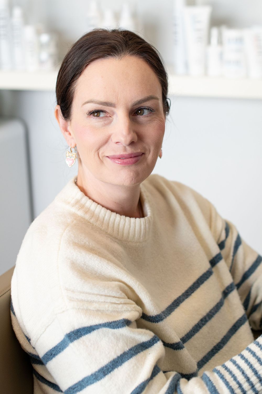 Woman in cream striped sweater smiling in a skincare studio with beauty products on shelves behind her.