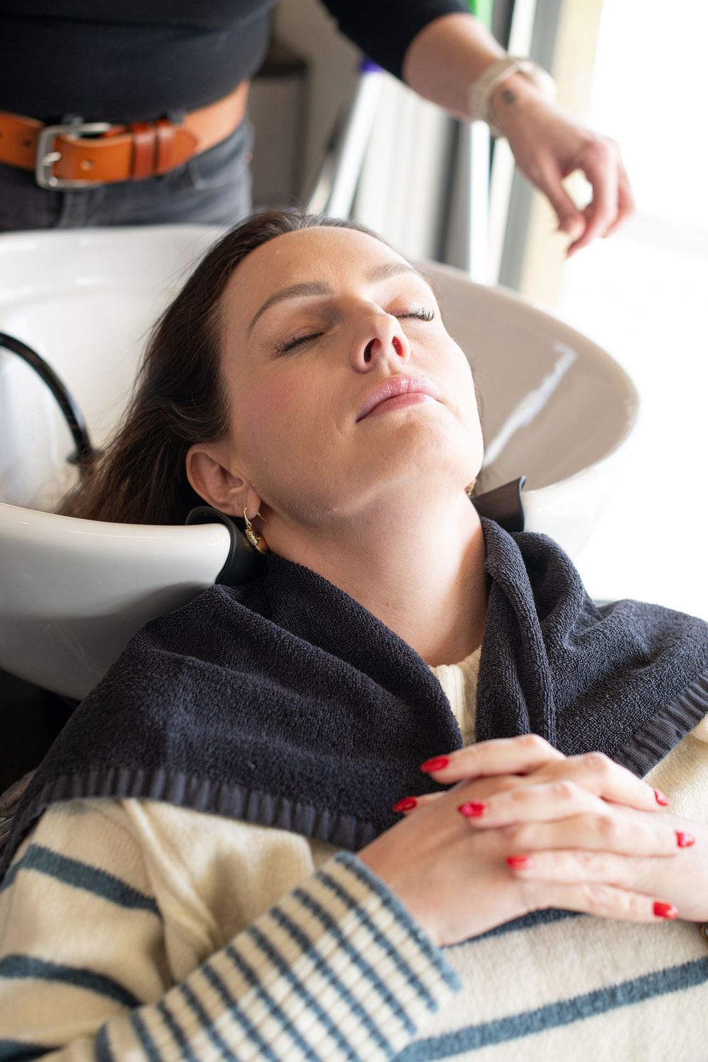 Woman relaxing at hair salon shampoo bowl with eyes closed, draped in towel, red nails folded on chest.