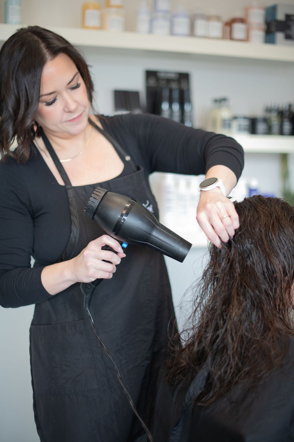 Female hairstylist blow drying long dark curly hair in a modern salon with products on shelves.