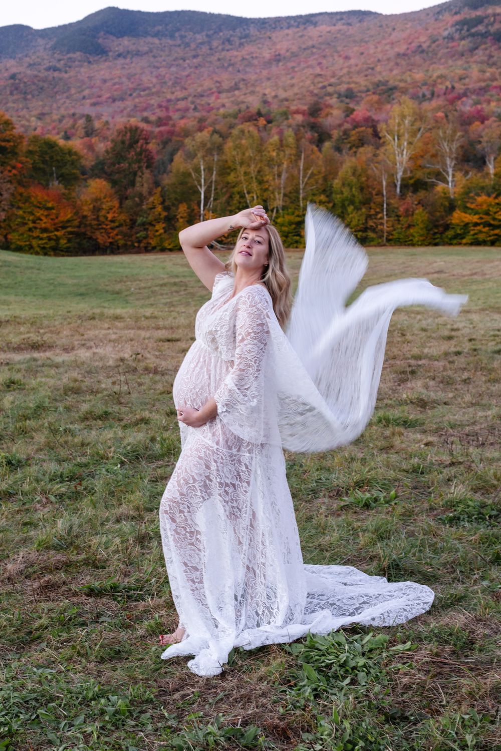 Expectant mother in white lace dress blows in breeze poses in open field with fall foliage. Stowe North Conway maternity photographer.