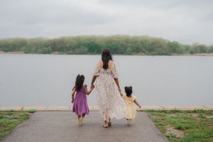 A mother in a floral dress holds hands with her two young daughters as they walk toward a calm lake on a cloudy day.