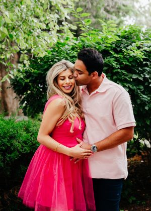 Smiling woman in a bright pink tulle dress embraced by a man in a pink polo kissing her cheek among green garden foliage.