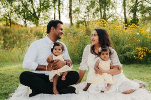 A smiling couple sits on a picnic blanket in a sunny meadow holding their two baby girls in matching cream dresses.