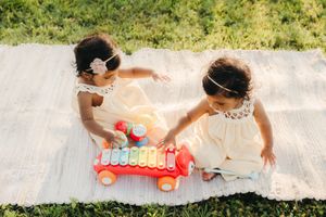 Two toddler girls in cream dresses and headbands play with a red toy xylophone car on a woven blanket in the grass.
