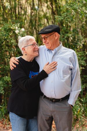 An older couple in glasses smiles warmly at each other while embracing in front of lush green bamboo during an outdoor portrait.