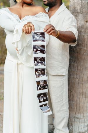 A pregnant woman and her partner hold a strip of ultrasound photos beside a tree, announcing their baby on the way.
