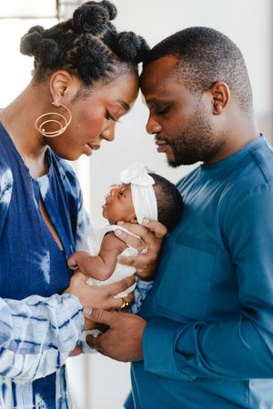 A loving couple touches foreheads while holding their newborn baby girl in a white bow headband during an intimate family portrait.