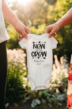 Two people hold up a white baby onesie reading New to the Crew, Baby Long 2024, in a sunlit garden for a pregnancy announcement.