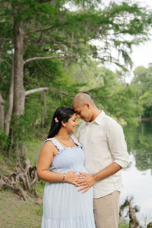 An expectant couple stands by a tranquil lake, touching foreheads as they gently cradle the baby bump during a maternity photo session.