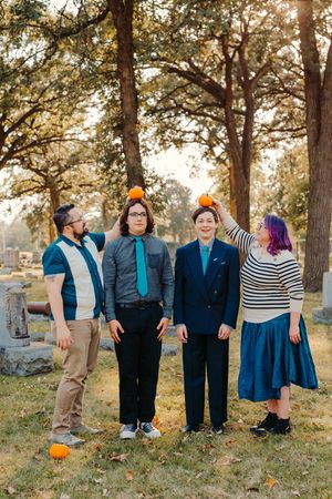 Four people pose in a cemetery, balancing small pumpkins on their heads, surrounded by autumn trees and fallen leaves.