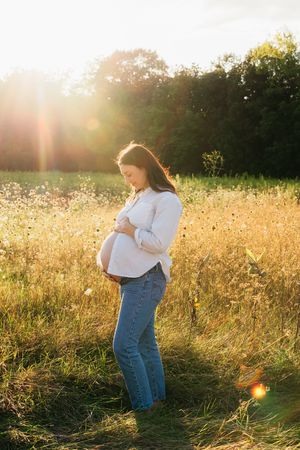 Pregnant woman in white shirt and jeans cradles her belly in a golden sunlit meadow during a maternity photo session.