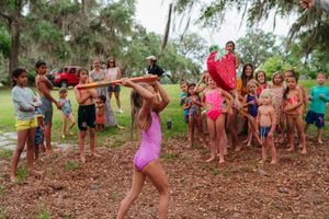 Girl in a purple swimsuit swings a stick at a piñata while a crowd of cheering children watches under shady trees.