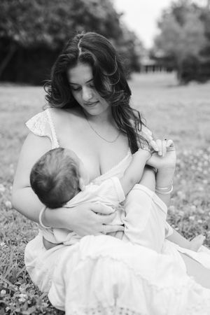 Black and white portrait of a mother in a white dress tenderly nursing her newborn baby while seated on a grassy lawn.
