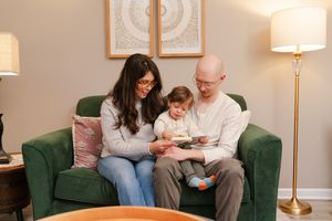 Mother and father read a picture book with their toddler on a green velvet sofa in a cozy, warmly lit living room.
