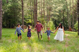 Family of five in a green forest clearing holds hands while walking toward the camera in a sunlit woodland setting.