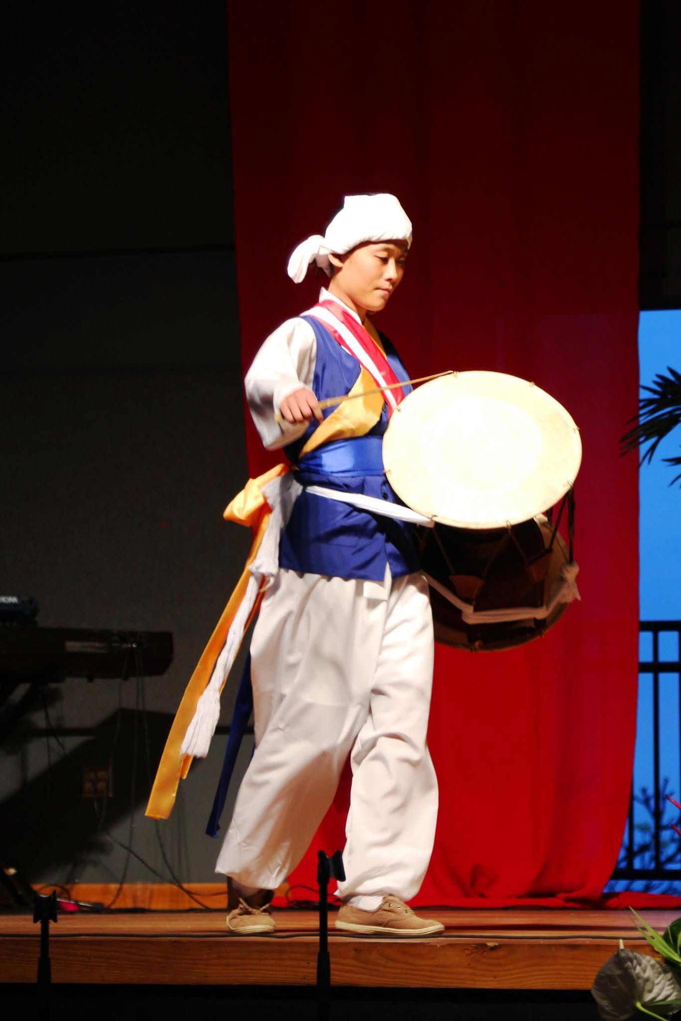 Performer in traditional Korean hanbok costume playing a janggu drum on stage during a cultural performance.