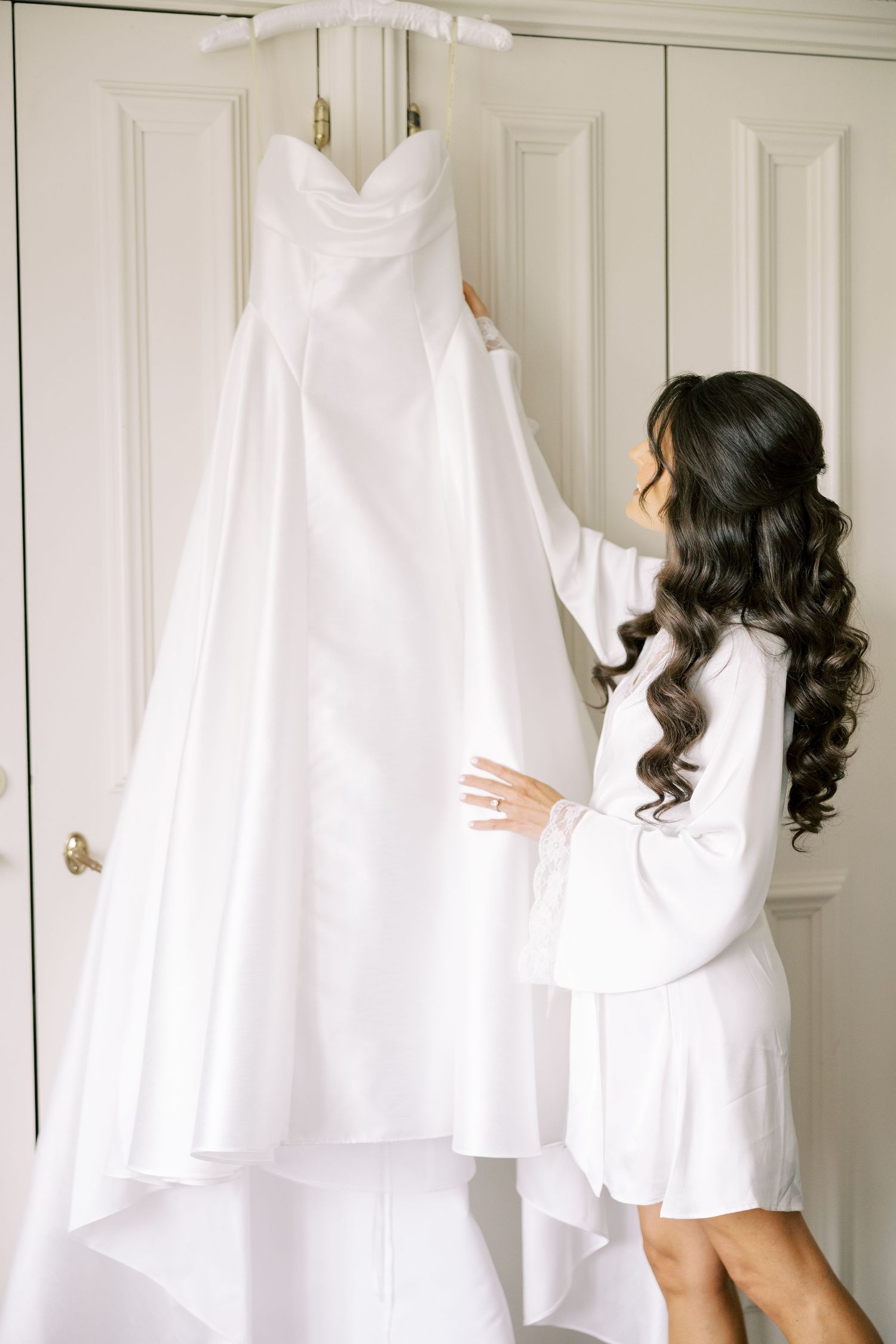 Bride in white robe admiring her elegant strapless wedding gown hanging on a wardrobe door.