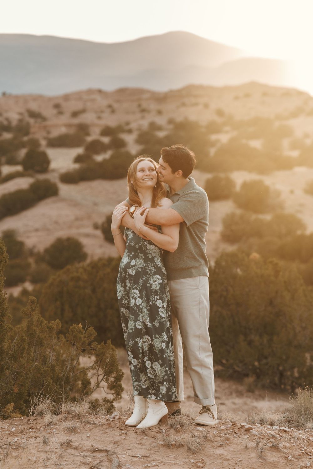 Couple embracing at golden hour in desert landscape, man kissing woman's cheek as mountains glow behind them.