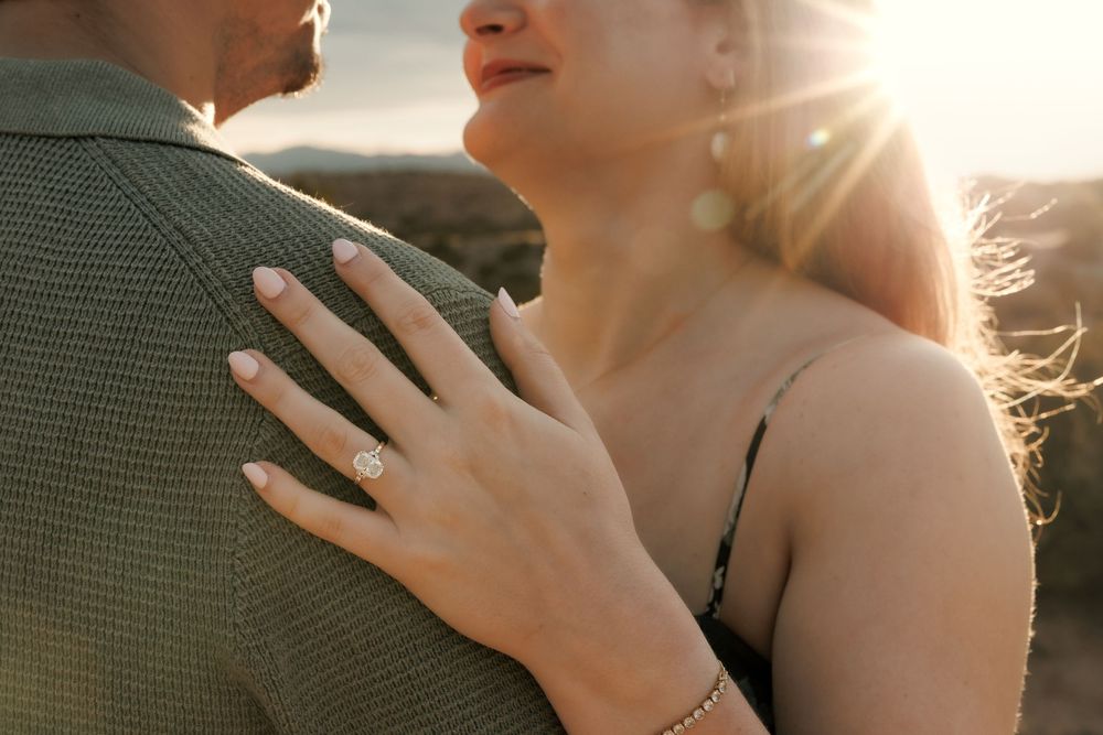 Couple embracing at sunset, woman wearing a diamond engagement ring, golden light glowing behind them.