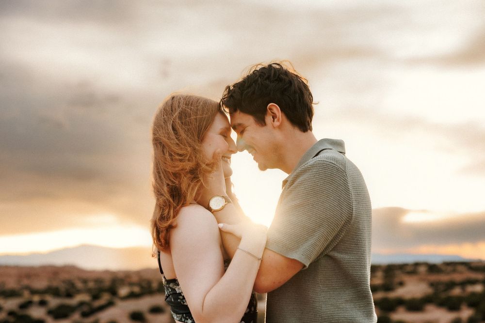Couple embracing at sunset in a desert landscape, touching foreheads romantically under dramatic cloudy skies.