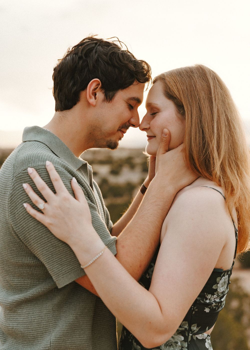 A romantic couple shares an intimate forehead-to-forehead moment at sunset, the woman in a floral dress and man in a sage polo.