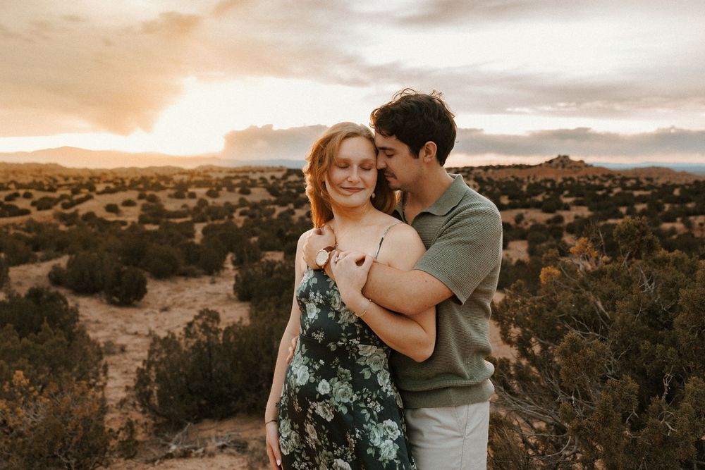 A couple embraces warmly at golden hour in a desert landscape, the woman in a floral dress smiling as her partner holds her close.