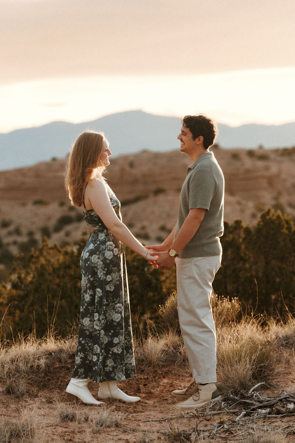 Smiling couple holding hands facing each other at sunset with mountain range backdrop in desert terrain.